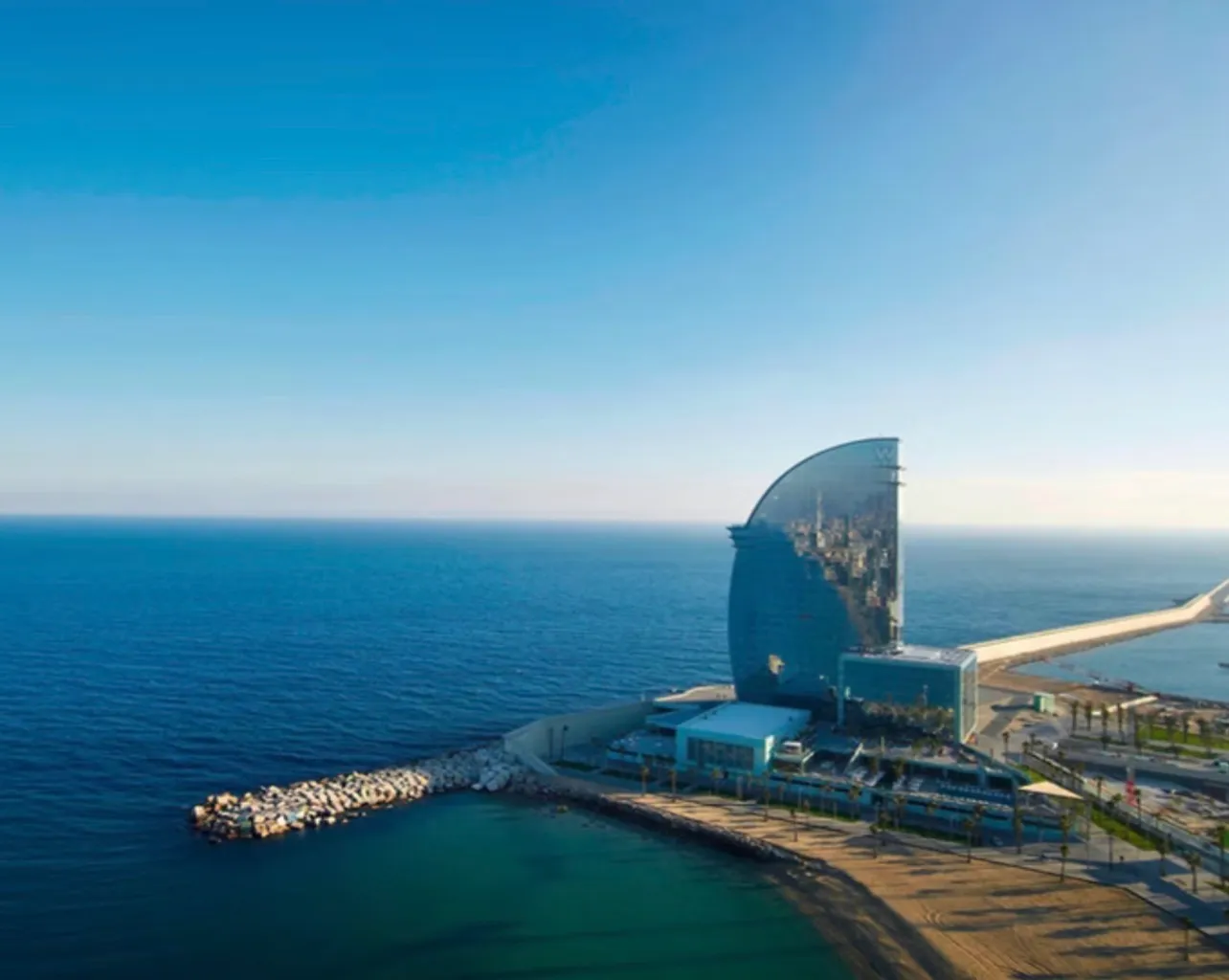 The hotel from the air, a sail-shaped tower standing at the end of the beach with a stone breakwater running out into the sea