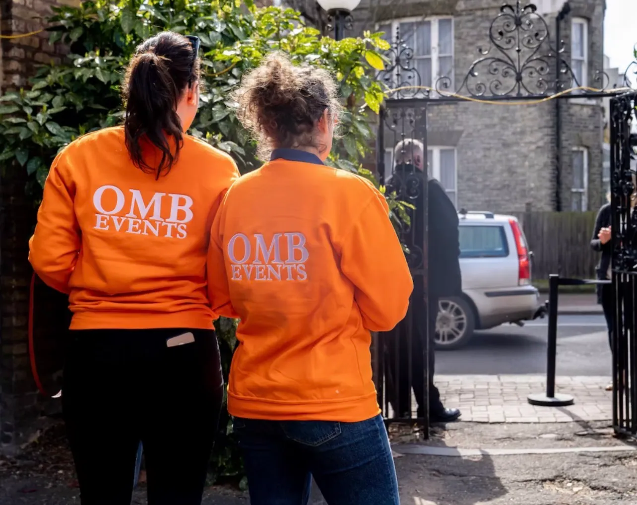 Two of the team in orange sweatshirts walking up to the club gate before it opens, a rope barrier set out on the pavement beyond it