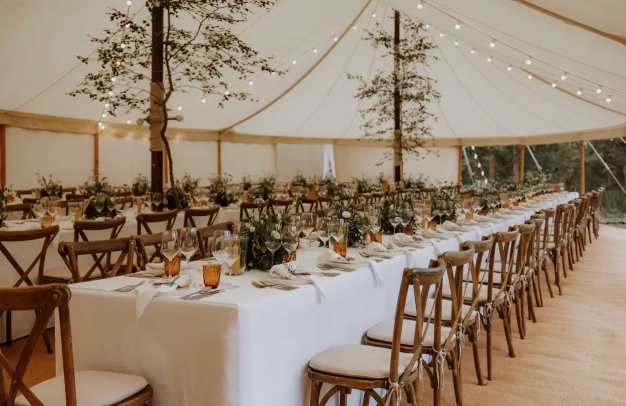 Long trestle tables dressed for a wedding breakfast under a pole marquee, foliage and candles down the centre and festoon lights strung overhead