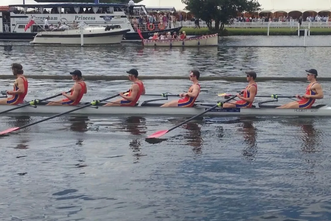 A crew racing past the enclosures, a launch and spectators on the far bank
