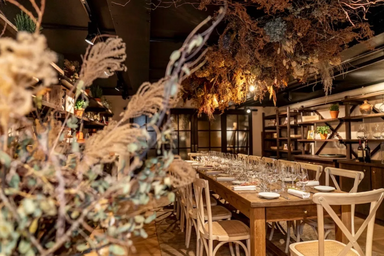 A long wooden table laid with glassware the length of it, under a ceiling installation of dried flowers and grasses lit from within