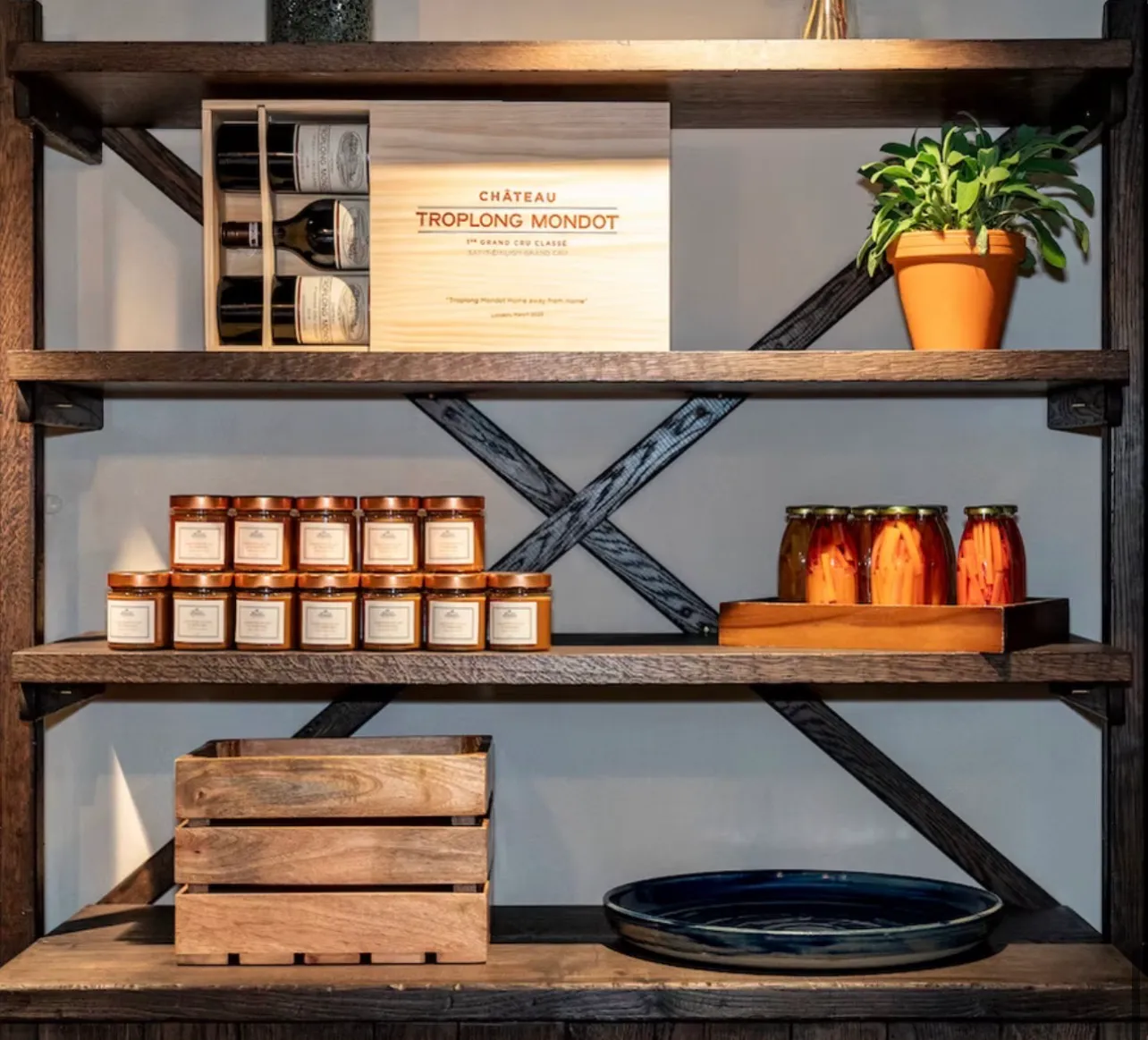A wooden wine case printed with the château's name standing on a shelf between potted herbs, jars of preserves and a stack of crates