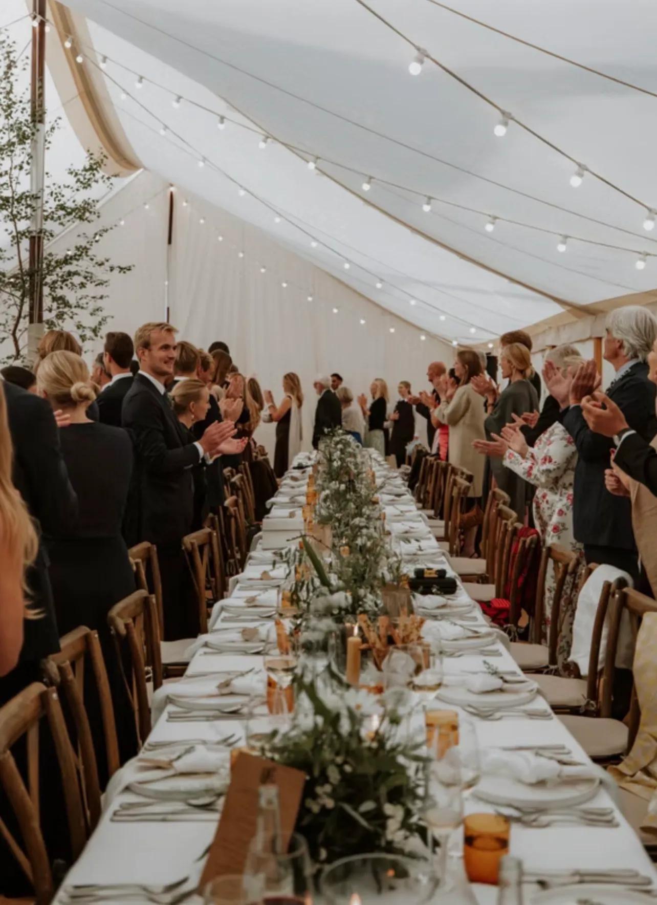 Guests standing and applauding along both sides of the long table under strings of festoon lights, places still laid in front of them