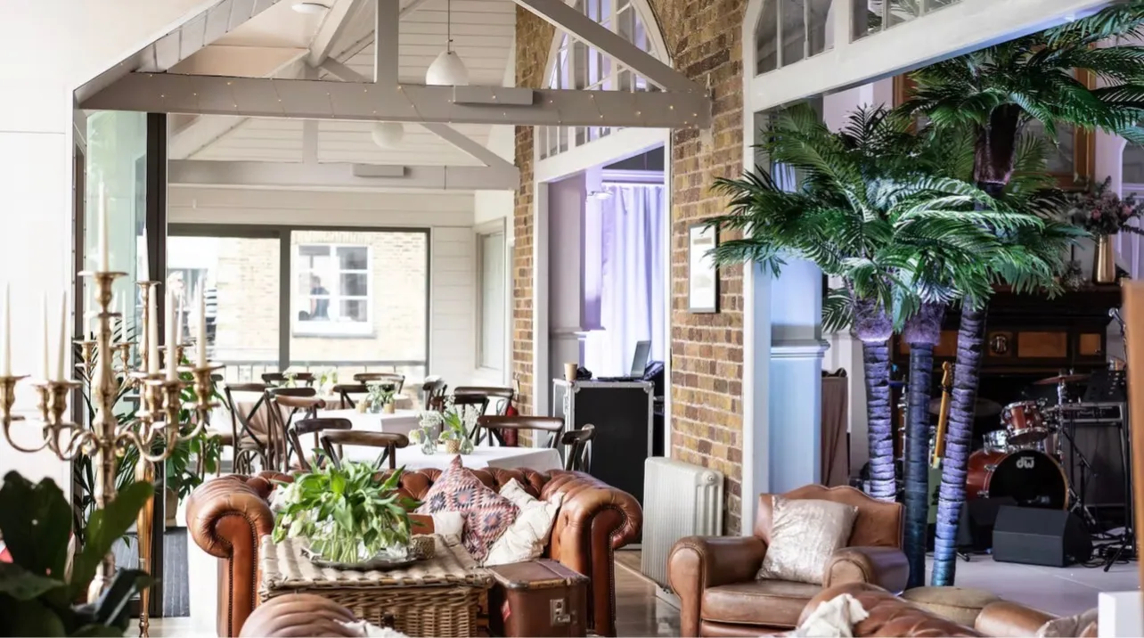 A club room dressed for an evening: dining tables laid beyond leather armchairs, with a stage and drum kit set up under the pitched roof